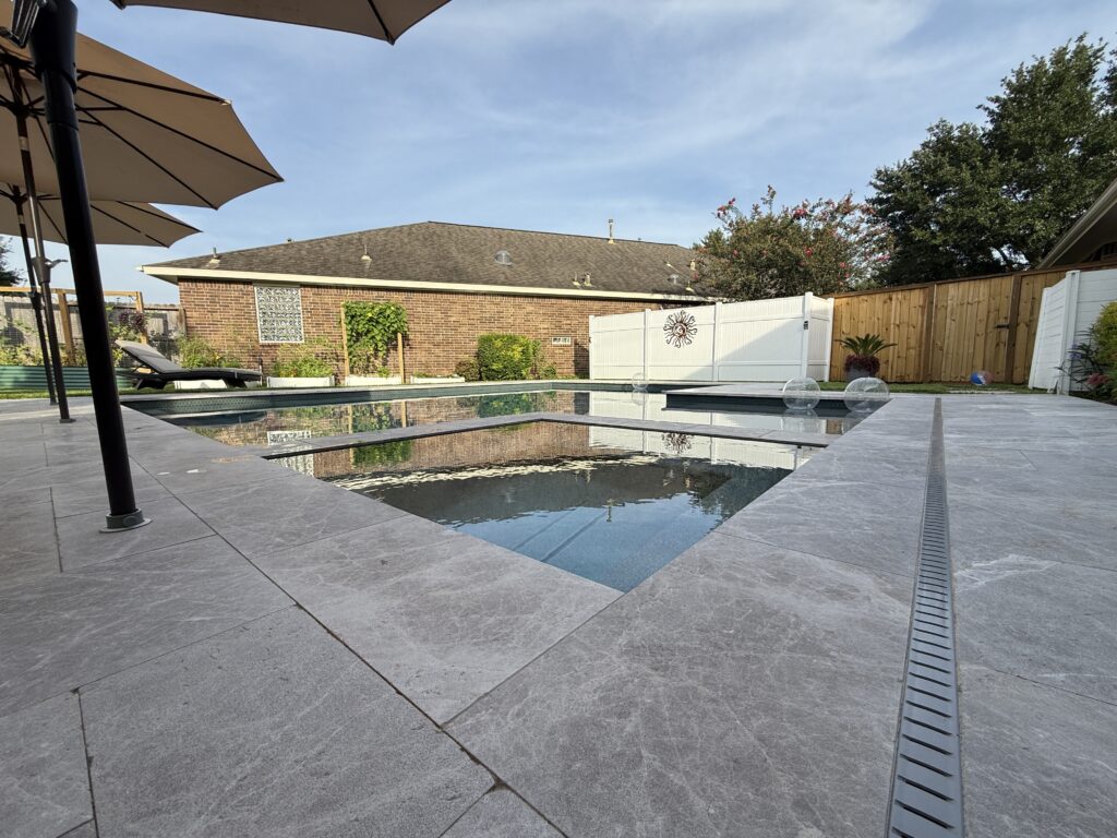Poolside backyard with grey stone paving, beige umbrellas, lounge chair, and a brick building in the background under a blue sky.