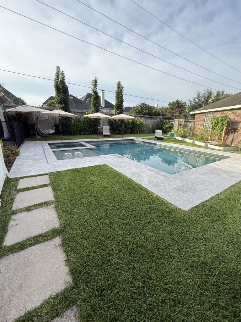 Backyard pool with stone patio, green lawn, and umbrellas over lounge chairs nearby.