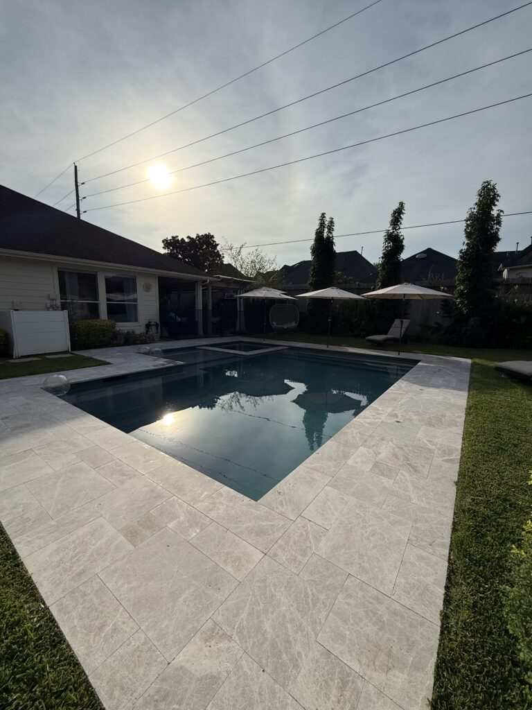 Residential backyard pool area with a rectangular blue pool, light stone decking, and white patio umbrellas. 2]