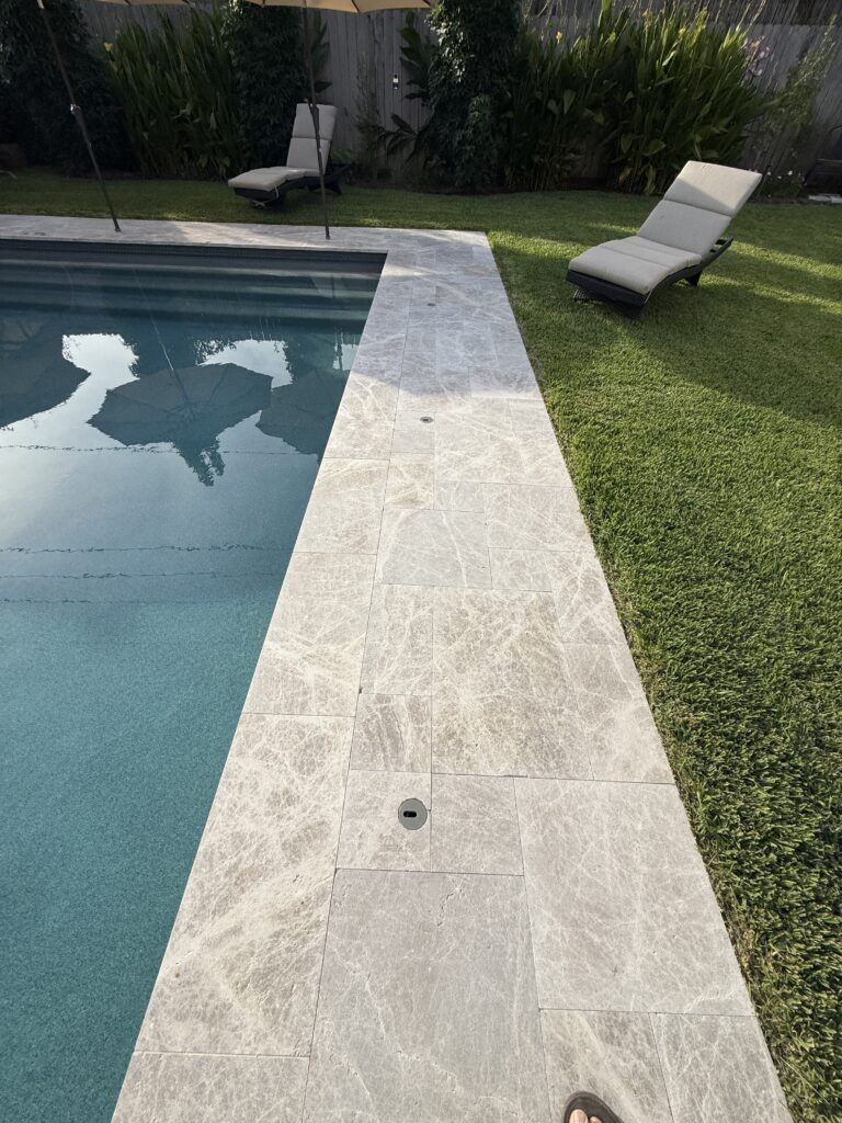 Poolside scene with a blue pool, light gray stone deck, and two beige cushioned lounge chairs; green lawn and plants in the background