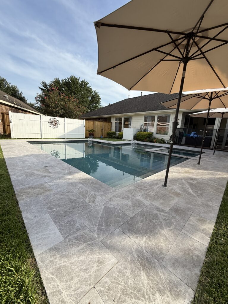 Rectangular in-ground pool with a gray stone deck and two large beige umbrellas in a sunny backyard.