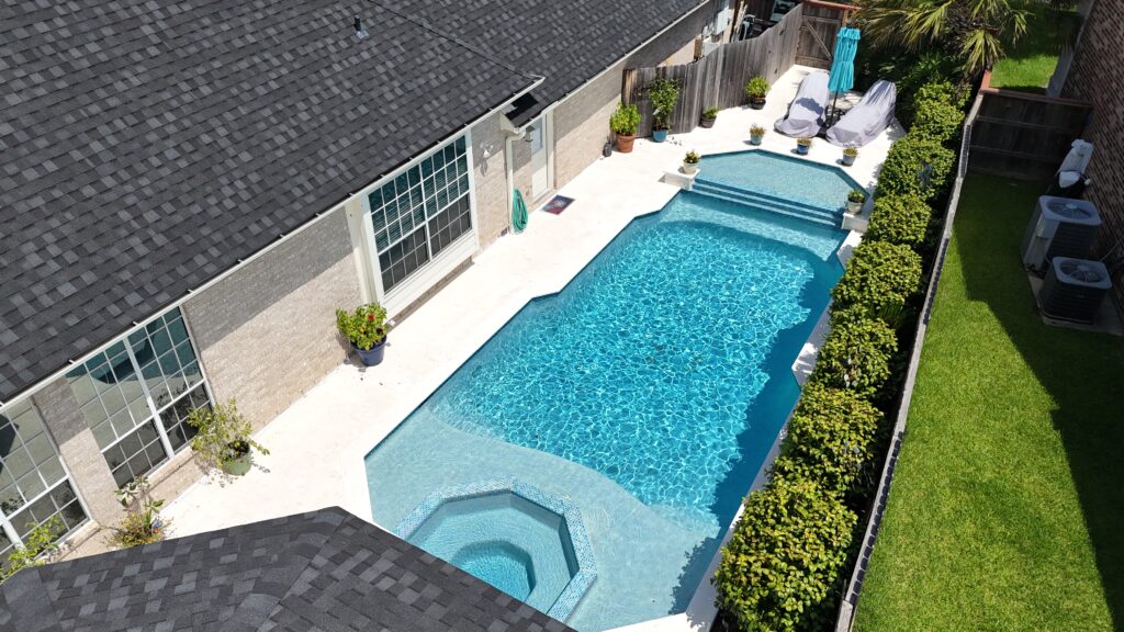 Aerial view of a suburban backyard pool with a connected spa and white concrete deck, potted plants, and patio furniture along a brick house wall.