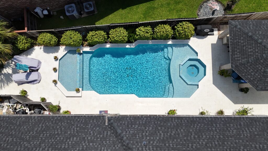 Aerial view of a backyard pool with a built-in spa, surrounded by a white deck and manicured shrubs along a fence.