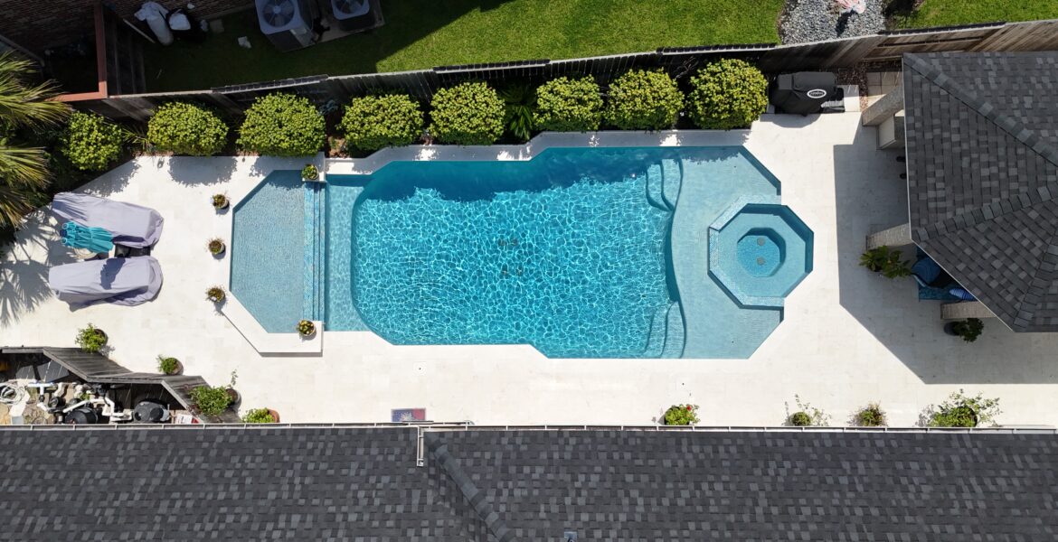 Aerial view of a backyard pool with a built-in spa, surrounded by a white deck and manicured shrubs along a fence.