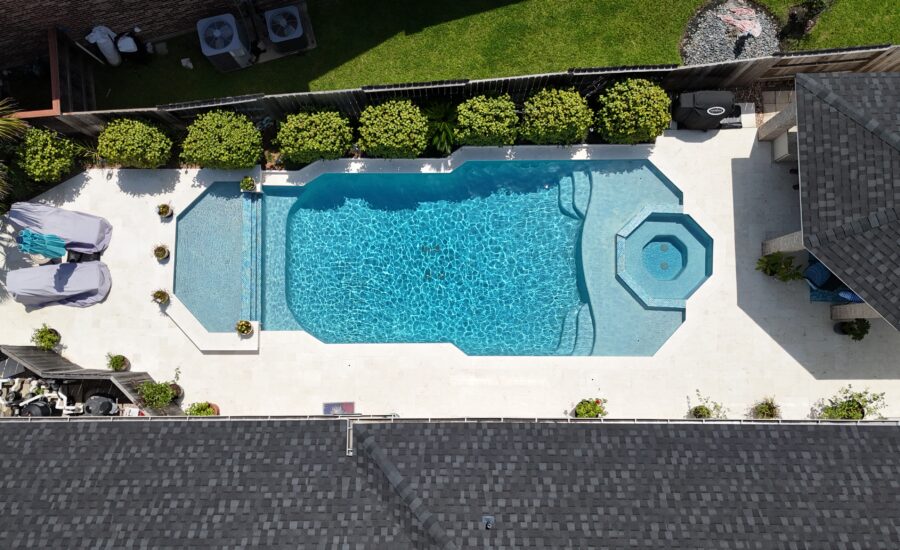 Aerial view of a backyard pool with a built-in spa, surrounded by a white deck and manicured shrubs along a fence.