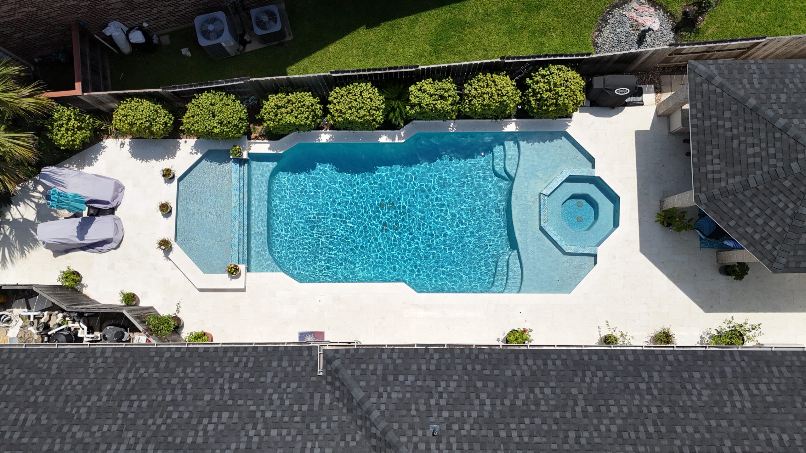 Aerial view of a backyard pool with a built-in spa, surrounded by a white deck and manicured shrubs along a fence.