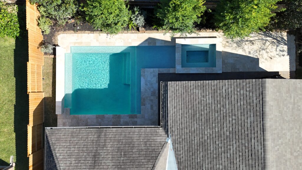 Aerial view of a modern backyard with a turquoise rectangular pool, built-in steps, and a square spa alcove surrounded by a stone patio and greenery.