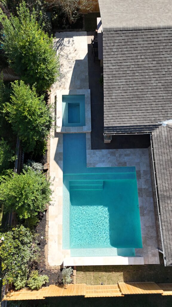 Aerial view of a backyard showing a rectangular turquoise pool, stone patio, and a grey-roofed house on the right surrounded by trees on the left.