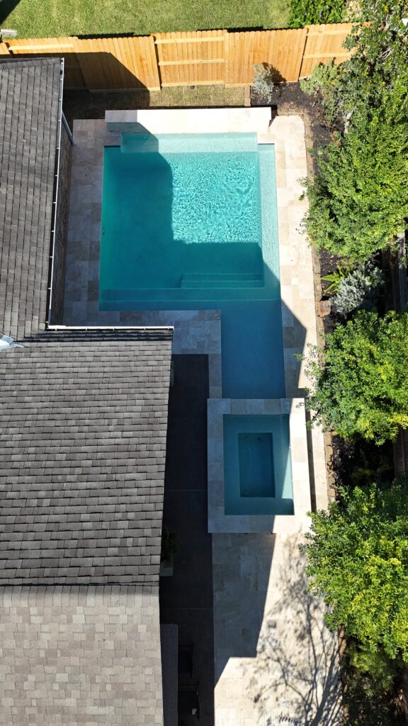 Aerial view of a rectangular turquoise pool with built-in steps, surrounded by stone patio, a wooden fence, and green shrubbery.