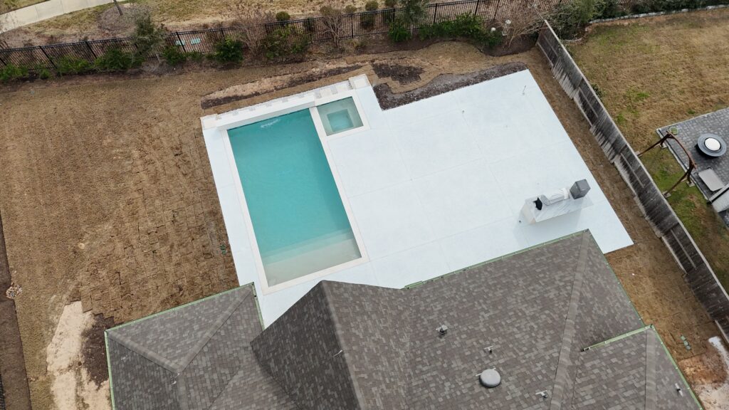 Aerial view of a modern backyard featuring a rectangular turquoise pool with a small square spa, surrounded by a white deck and brown dirt yard, fenced perimeter.