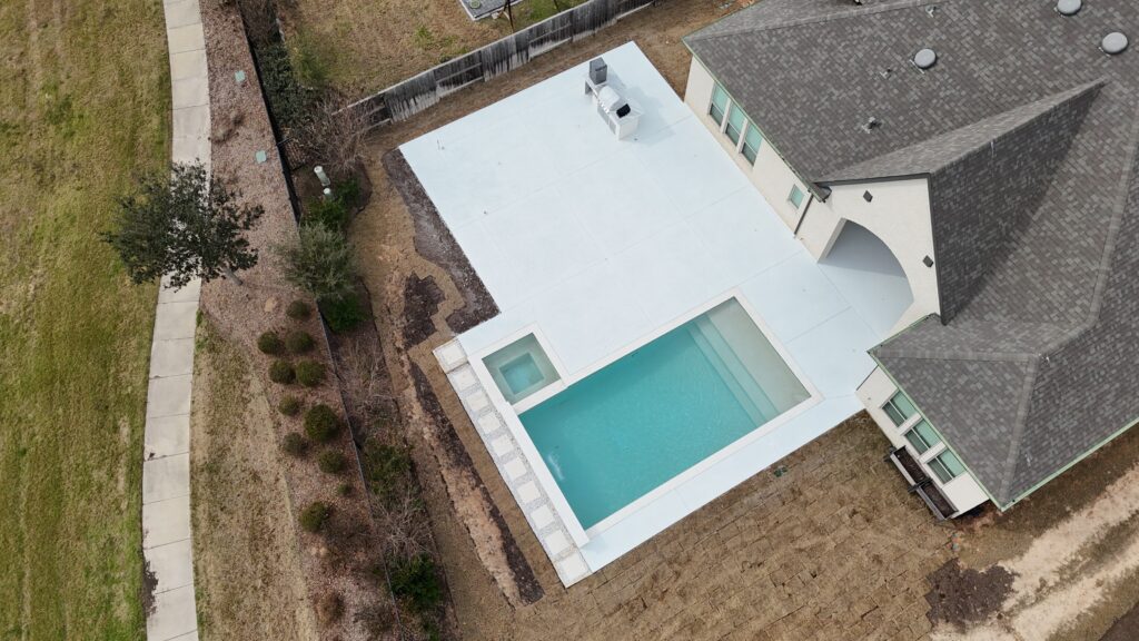 Aerial view of a house with a light blue rectangular pool in the backyard, surrounded by a white concrete deck and a gray shingle roof.