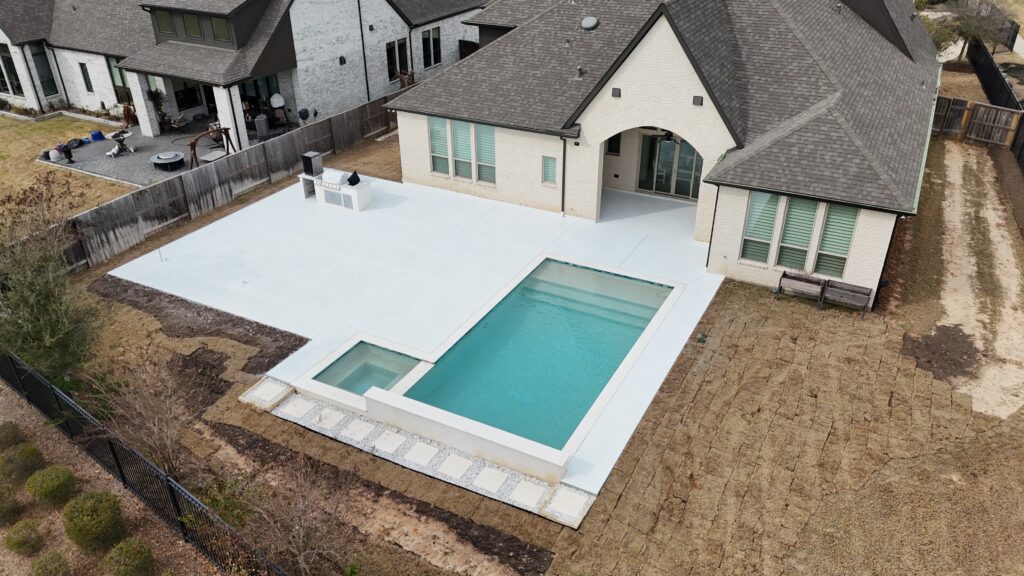 Aerial view of a backyard with a rectangular pool and a small spa, surrounded by a white concrete deck and a beige brick house.