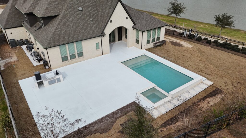 Aerial view of a modern beige brick house with a gray roof, large white concrete patio, and a rectangular pool with a spa at the edge of a fenced yard by a lake.