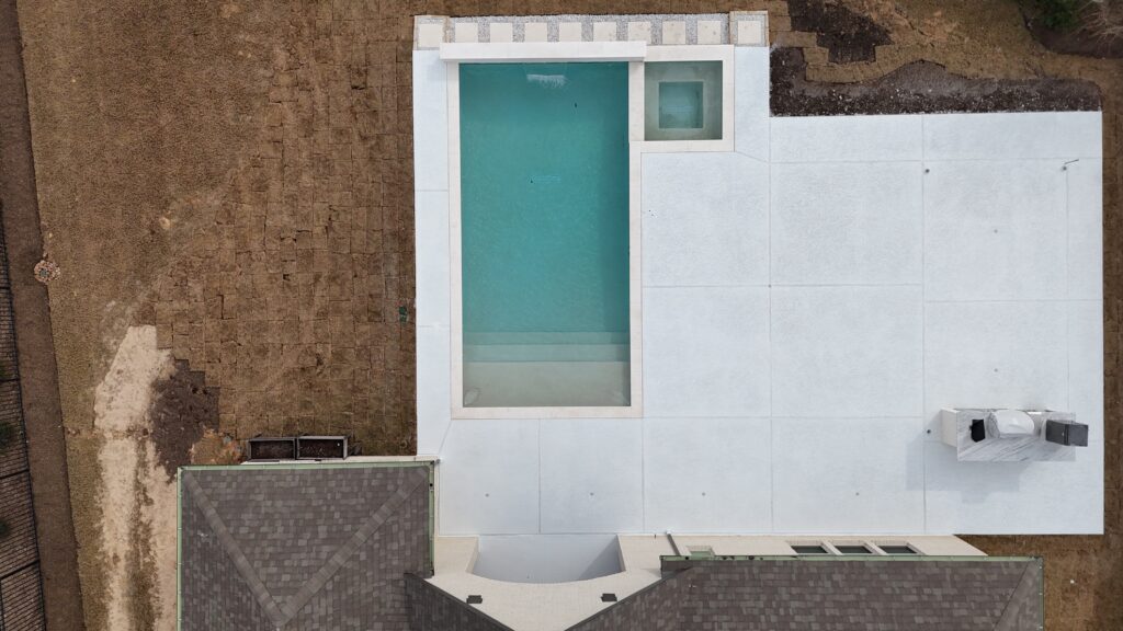 Aerial view of a modern building rooftop featuring a turquoise rectangular pool bordered by light stone, with a small square spa area to the right and expansive light-gray tile surfaces; brown dirt ground and a neighboring roof frame the scene.
