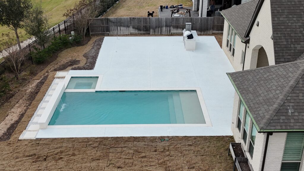 Aerial view of a modern backyard: rectangular pool with a white cover on a pale-blue deck, brick house on the right, wooden fence in the back, and a patio with people and dogs in the distance.