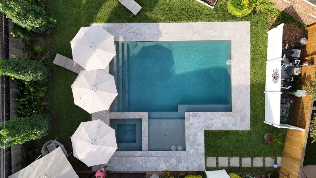 Aerial view of a rectangular swimming pool surrounded by light stone patio, white umbrellas, lounge chairs, and green lawn nearby.