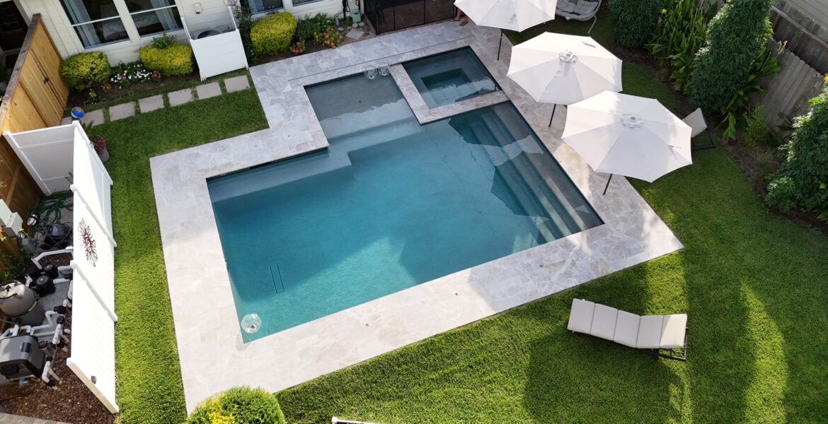 Aerial view of a modern backyard pool area with a large rectangular pool, a shallow step area, and a smaller attached spa, surrounded by light stone decking and green lawn under white patio umbrellas and lounge chairs.