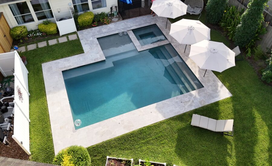 Aerial view of a modern backyard pool area with a large rectangular pool, a shallow step area, and a smaller attached spa, surrounded by light stone decking and green lawn under white patio umbrellas and lounge chairs.