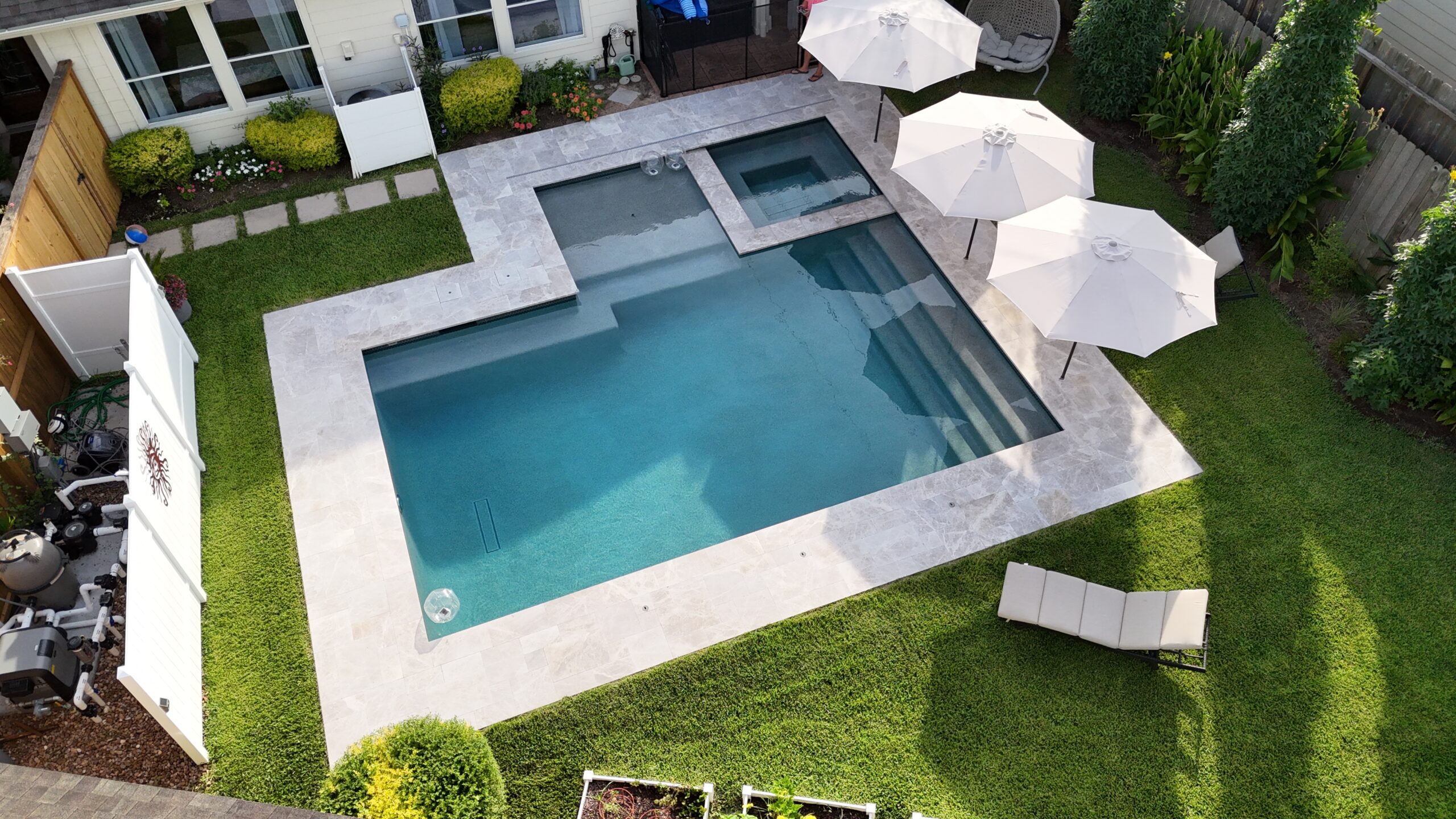 Aerial view of a modern backyard pool area with a large rectangular pool, a shallow step area, and a smaller attached spa, surrounded by light stone decking and green lawn under white patio umbrellas and lounge chairs.