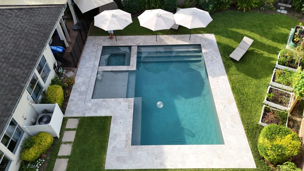 Aerial view of a modern rectangular pool with gray stone deck, white umbrellas, and a lounge chair in a green backyard.