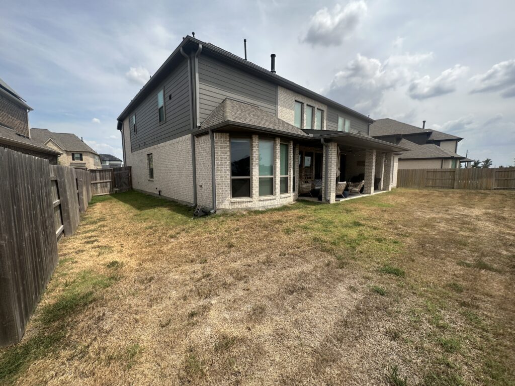 Backyard of a suburban home showing brick and gray siding, covered patio with chairs, wooden fence, and a cloudy sky above.