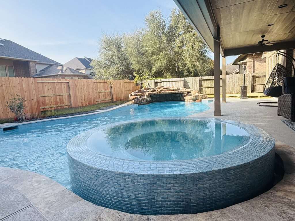 Backyard pool with a raised circular spa, stone waterfall, wooden fence, and covered patio on the right.