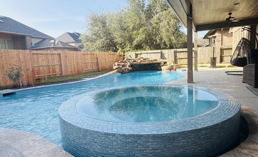 Backyard pool with a raised circular spa, stone waterfall, wooden fence, and covered patio on the right.