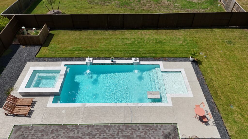 Aerial drone view of a custom rectangular pool and raised spa with tanning ledge, wide concrete coping, and decorative gravel border — completed by Beach'n Pools in Cane Island, Katy TX