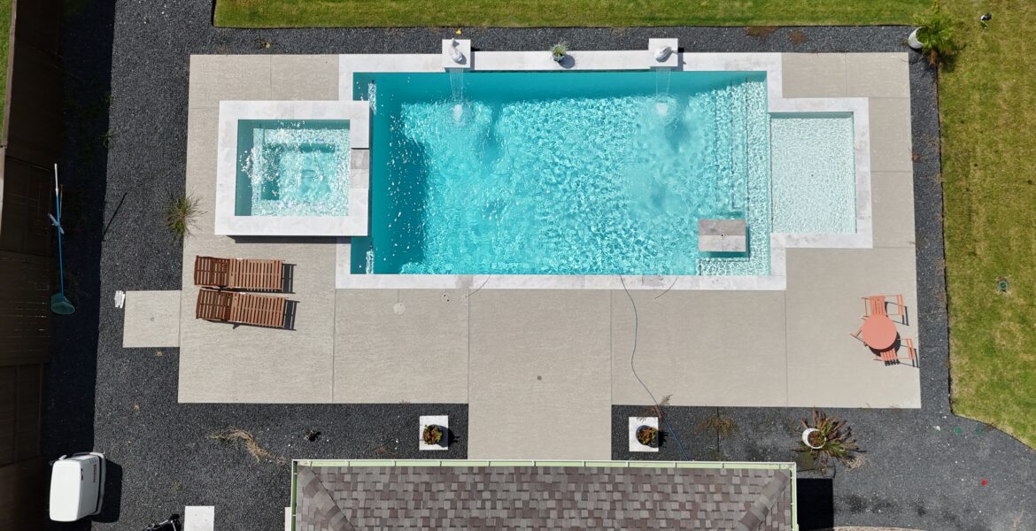 Aerial view of a rectangular swimming pool with a built-in spa, surrounded by light-gray concrete patio and gravel, with lounge chairs and a small table on the deck and green lawn around.