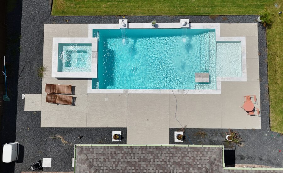 Aerial view of a rectangular swimming pool with a built-in spa, surrounded by light-gray concrete patio and gravel, with lounge chairs and a small table on the deck and green lawn around.
