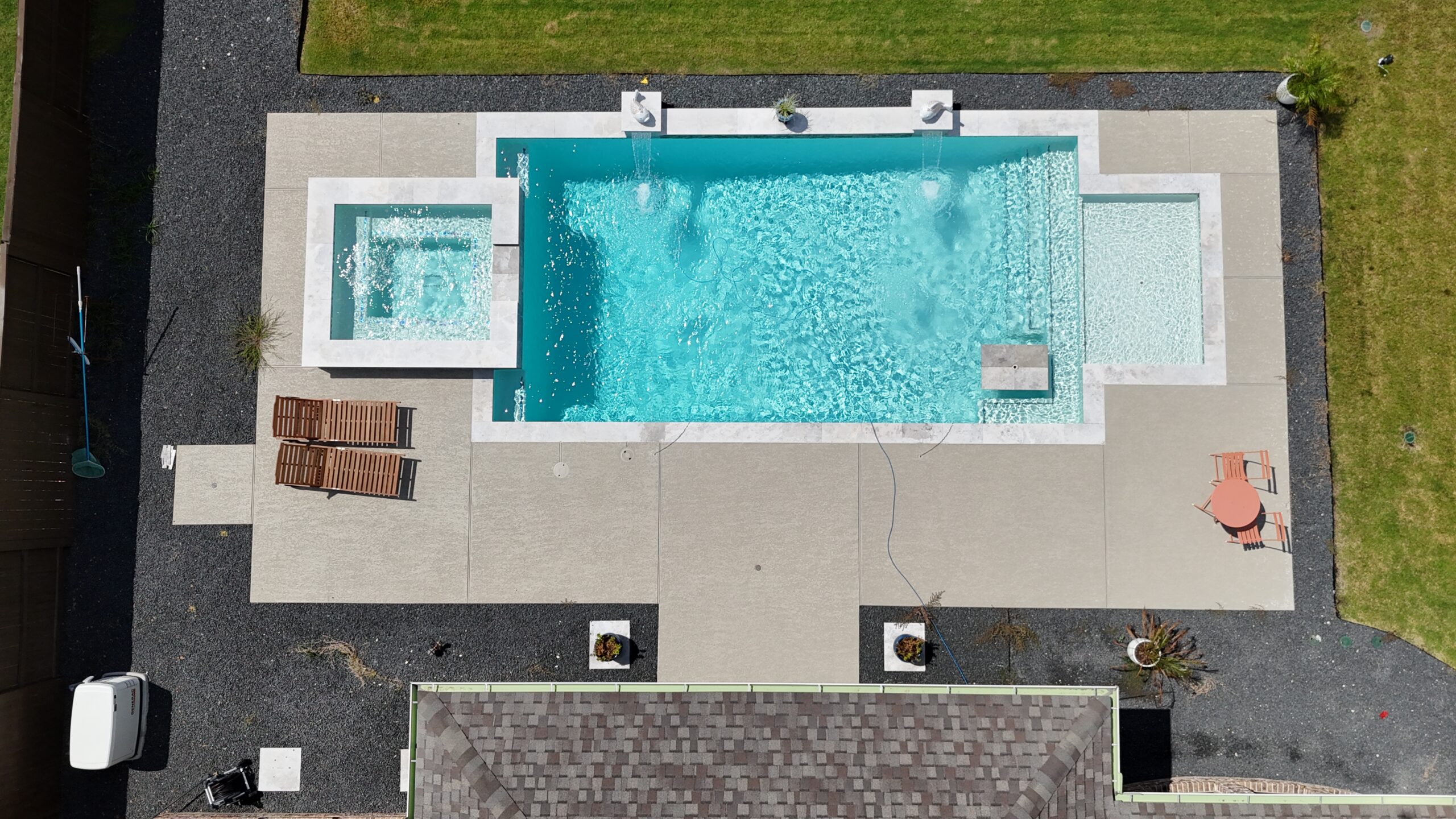 Aerial view of a rectangular swimming pool with a built-in spa, surrounded by light-gray concrete patio and gravel, with lounge chairs and a small table on the deck and green lawn around.