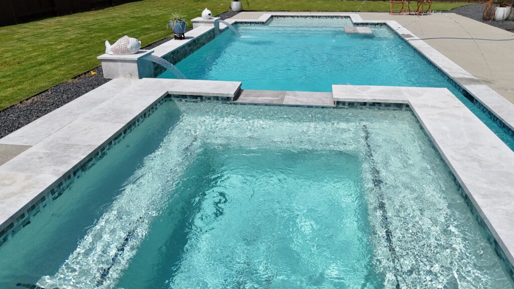 Aerial drone view of a custom rectangular pool and raised spa with tanning ledge, wide concrete coping, and decorative gravel border — completed by Beach'n Pools in Cane Island, Katy TX
