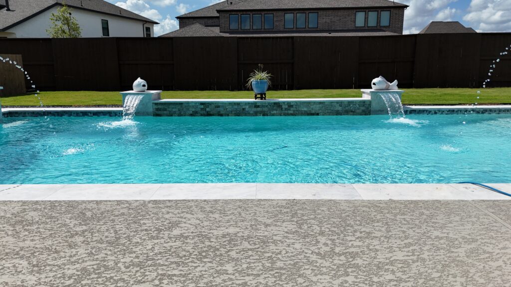 Aerial drone view of a custom rectangular pool and raised spa with tanning ledge, wide concrete coping, and decorative gravel border — completed by Beach'n Pools in Cane Island, Katy TX