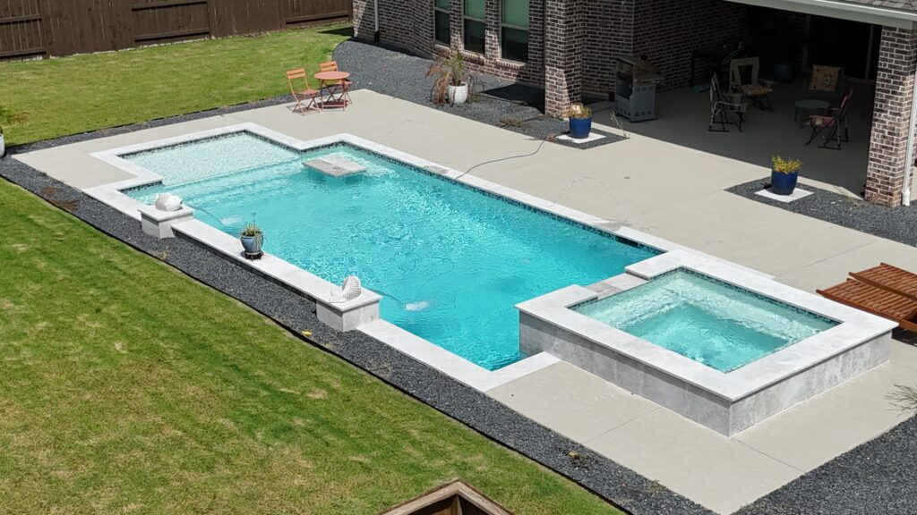 Aerial drone view of a custom rectangular pool and raised spa with tanning ledge, wide concrete coping, and decorative gravel border — completed by Beach'n Pools in Cane Island, Katy TX