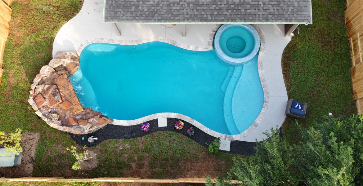 Aerial drone view of a custom freeform inground pool with attached circular spa and natural rock waterfall, surrounded by lush green landscaping in Katy, TX, built by Beach'n Pools