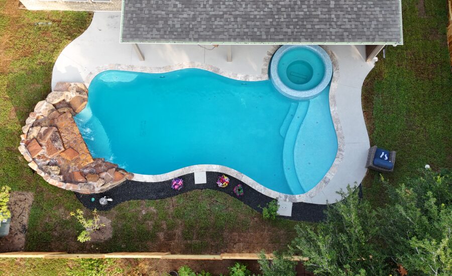 Aerial drone view of a custom freeform inground pool with attached circular spa and natural rock waterfall, surrounded by lush green landscaping in Katy, TX, built by Beach'n Pools