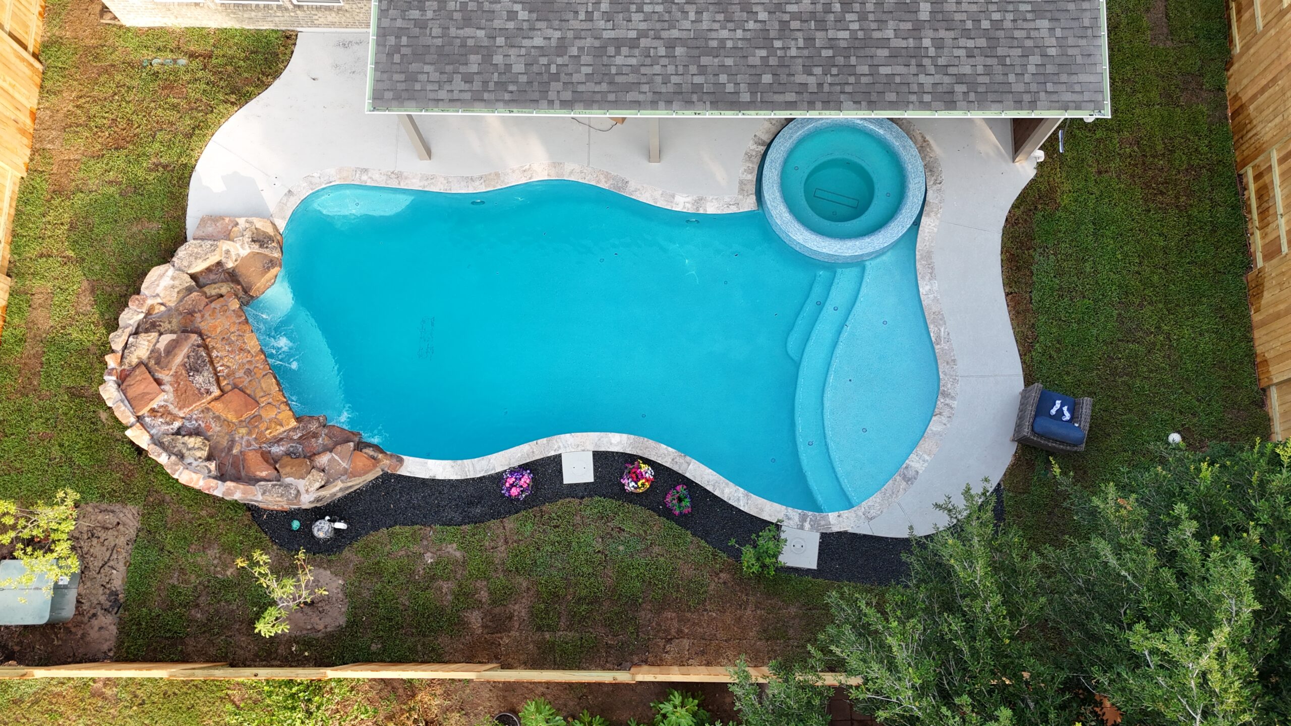 Aerial drone view of a custom freeform inground pool with attached circular spa and natural rock waterfall, surrounded by lush green landscaping in Katy, TX, built by Beach'n Pools